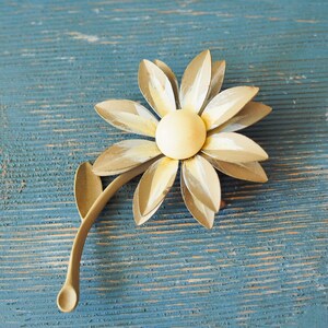 K&ouml;nnte beinhalten: Eine goldene Brosche in Form einer Blume mit gelbem Zentrum und wei&szlig;en Akzenten. Die Blume hat einen Stiel mit einem Blatt.