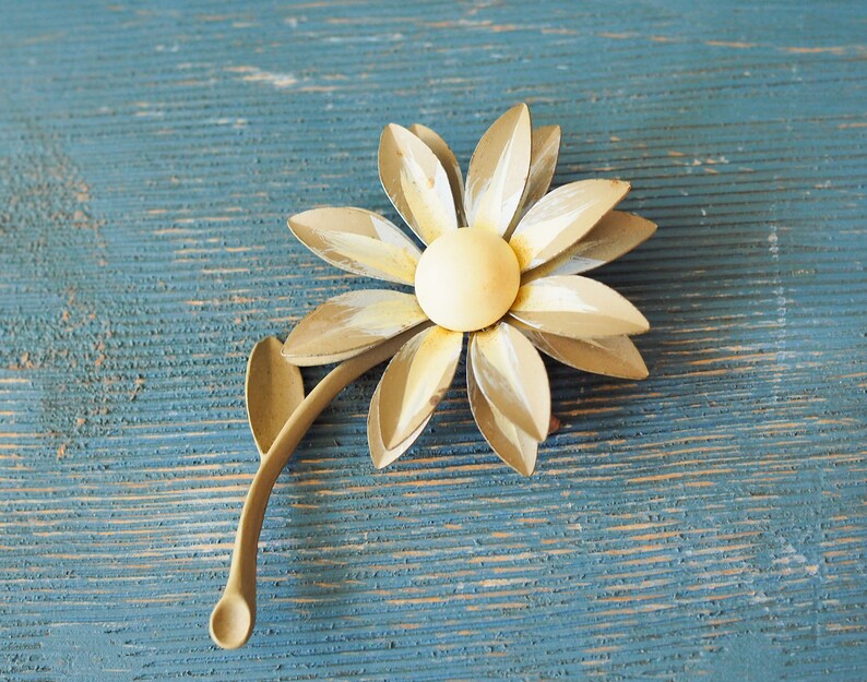 K&ouml;nnte beinhalten: Eine goldene Brosche in Form einer Blume mit gelbem Zentrum und wei&szlig;en Akzenten. Die Blume hat einen Stiel mit einem Blatt.