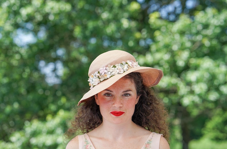 May include: A woman with curly brown hair wears a light brown hat with a white flower trim. The hat is tilted at an angle, and the woman is looking directly at the camera.