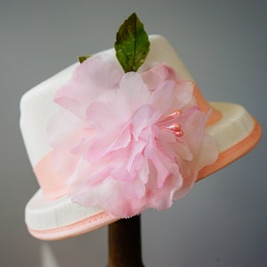 Op de afbeelding: Een witte hoed met een roze bloem en een roze lint.