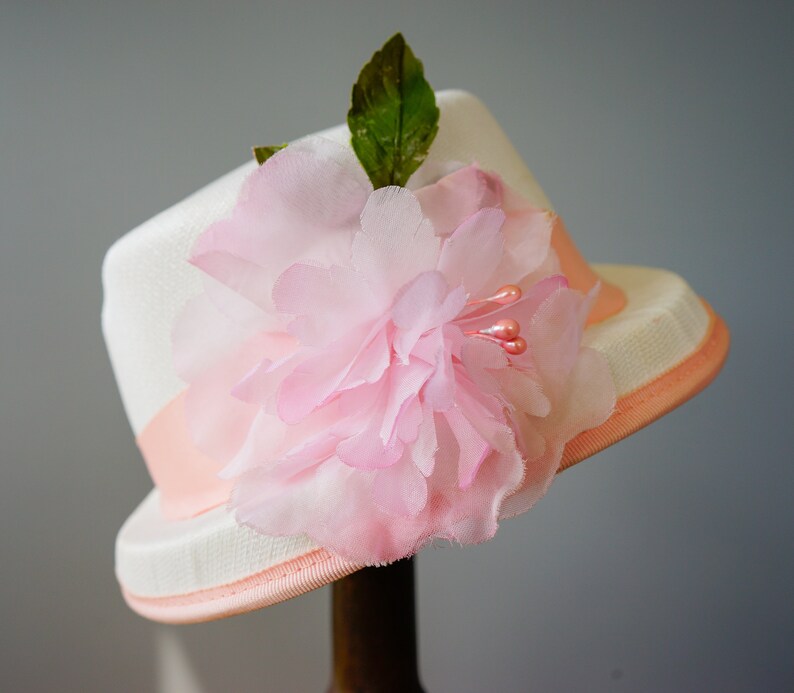 Op de afbeelding: Een witte hoed met een roze bloem en een roze lint.