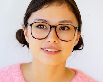 May include: A young person with dark hair wearing tortoiseshell glasses and a pink knitted sweater.