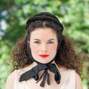 May include: A woman with curly dark hair is wearing a black bonnet with a black ribbon tied at the neck. She is wearing a light pink blouse with a floral pattern.