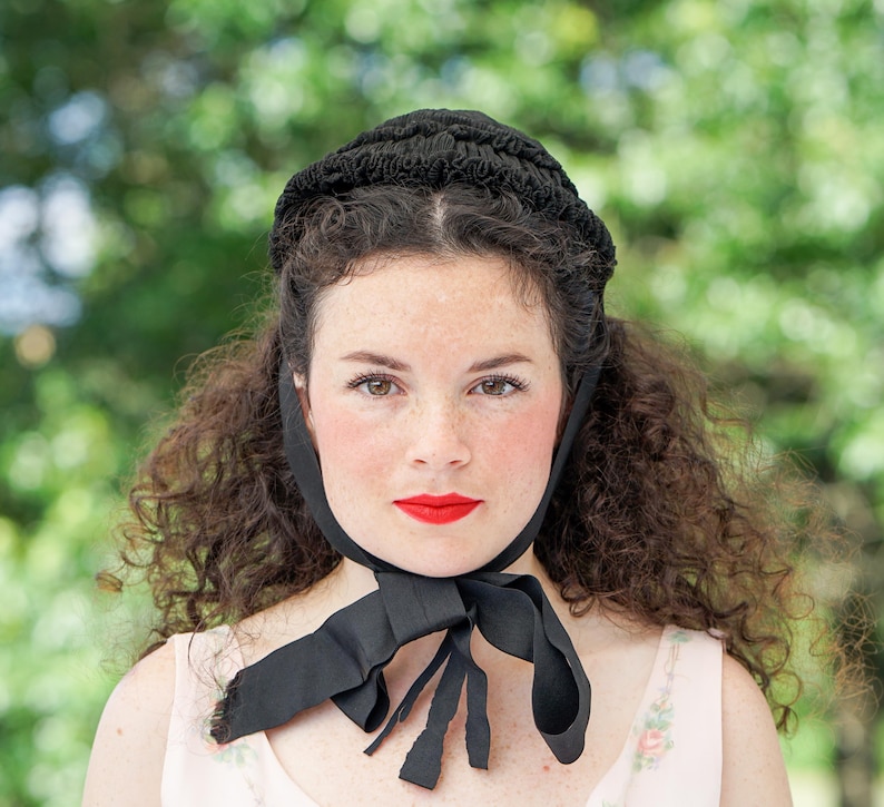 May include: A woman with curly dark hair is wearing a black bonnet with a black ribbon tied at the neck. She is wearing a light pink blouse with a floral pattern.