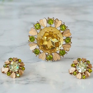 Peut inclure: Un ensemble de bijoux en or avec une grosse broche en strass jaune et deux boucles d'oreilles assorties. La broche est entour&eacute;e de petits strass verts et de feuilles dor&eacute;es. Les boucles d'oreilles sont &eacute;galement entour&eacute;es de petits strass verts et de feuilles dor&eacute;es.