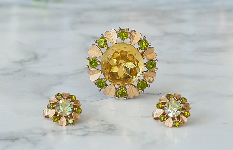 Peut inclure: Un ensemble de bijoux en or avec une grosse broche en strass jaune et deux boucles d'oreilles assorties. La broche est entour&eacute;e de petits strass verts et de feuilles dor&eacute;es. Les boucles d'oreilles sont &eacute;galement entour&eacute;es de petits strass verts et de feuilles dor&eacute;es.