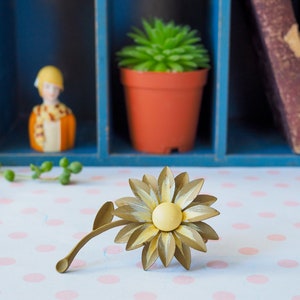 K&ouml;nnte beinhalten: Eine goldfarbene Metall-Sonnenblumenbrosche mit gelbem Zentrum und Stiel. Die Brosche liegt auf einer wei&szlig;en Oberfl&auml;che mit rosa Punkten.