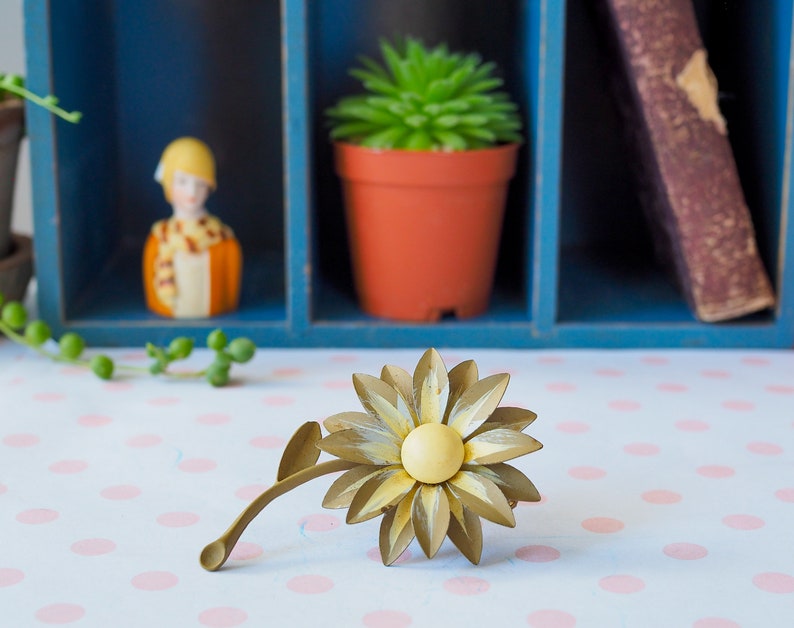 K&ouml;nnte beinhalten: Eine goldfarbene Metall-Sonnenblumenbrosche mit gelbem Zentrum und Stiel. Die Brosche liegt auf einer wei&szlig;en Oberfl&auml;che mit rosa Punkten.