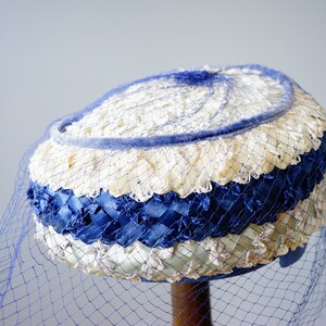 May include: A vintage hat with a blue veil. The hat has a woven design with alternating bands of white, blue, and cream-coloured materials. A blue net veil drapes over the hat, adding a touch of elegance. The hat is displayed on a wooden stand.