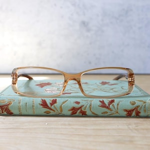 May include: A pair of brown-framed glasses with a light blue book with floral designs and the text "L'autre-Mer" on the cover.