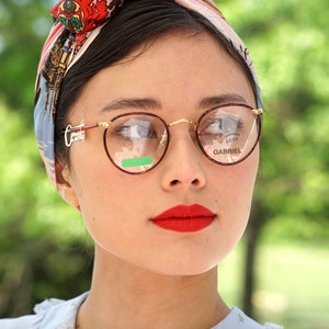 May include: A woman wearing a floral patterned headscarf and round, gold-rimmed glasses. The glasses have the words "GABRIEL" and "OPTICAL LENS" printed on the lenses.