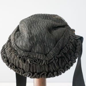 May include: A black fabric hat with a ruffled trim. The hat is made of a pleated fabric and has a wide brim.