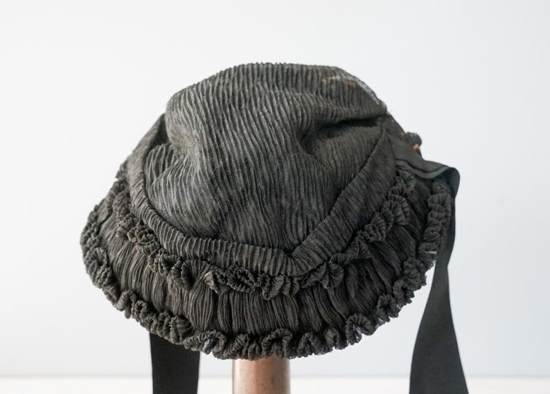 May include: A black fabric hat with a ruffled trim. The hat is made of a pleated fabric and has a wide brim.