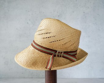 May include: A straw hat with a wide brim and a decorative band. The hat is made of woven straw and features a brown, red, and green striped band around the crown. The hat has a classic fedora shape.