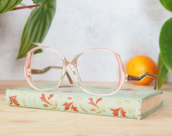 May include: Clear pink plastic framed glasses with a rounded square shape. The glasses are resting on a green book with gold lettering.