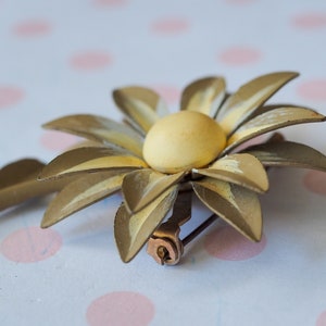 K&ouml;nnte beinhalten: Eine goldfarbene Brosche aus Metall in Form einer Blume mit gelbem Zentrum. Die Blume hat eine Nadel auf der R&uuml;ckseite zum Anbringen an Kleidung.