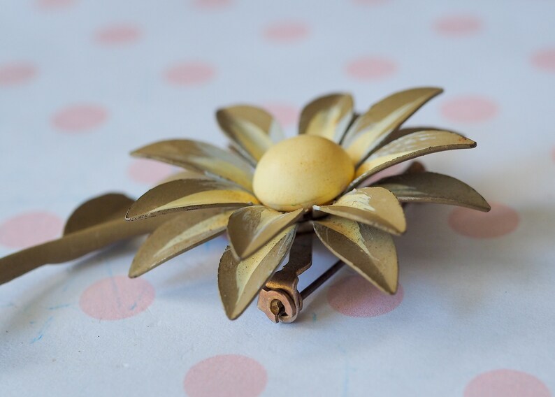 K&ouml;nnte beinhalten: Eine goldfarbene Brosche aus Metall in Form einer Blume mit gelbem Zentrum. Die Blume hat eine Nadel auf der R&uuml;ckseite zum Anbringen an Kleidung.