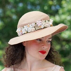 May include: A wide-brimmed straw hat with a band of white and pink fabric flowers. The hat is beige and has a slightly rounded crown.
