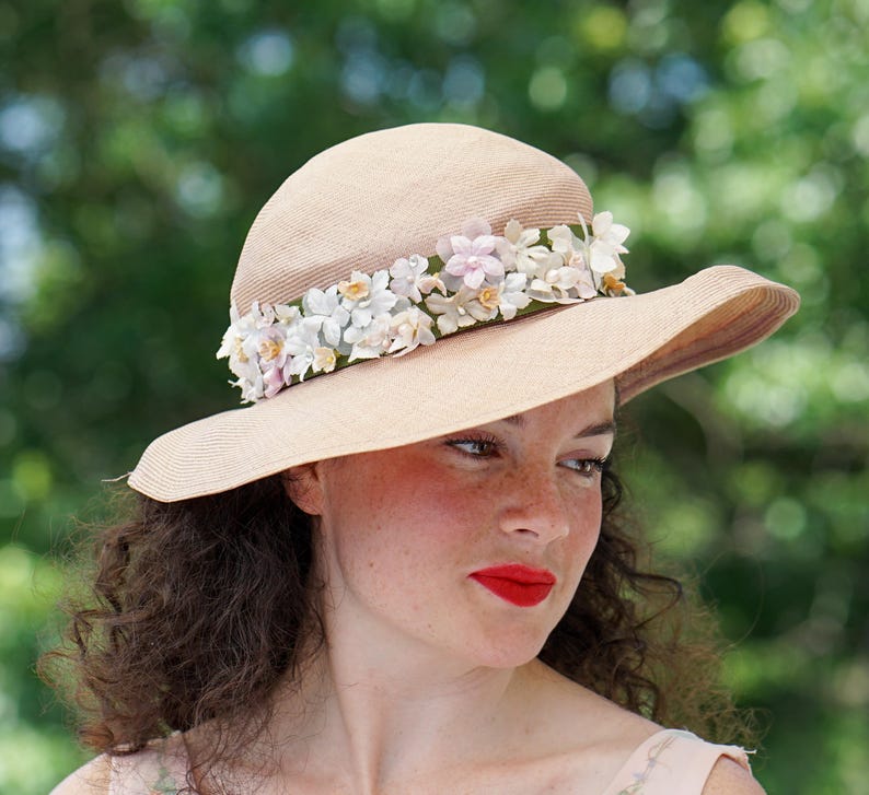 May include: A wide-brimmed straw hat with a band of white and pink fabric flowers. The hat is beige and has a slightly rounded crown.