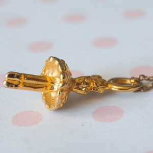 May include: A gold-colored pendant necklace featuring a detailed ballerina figure. The ballerina is in a classical pose, with arms extended. The necklace chain is delicate and complements the pendant's intricate design. The background is a soft, blurred pattern.