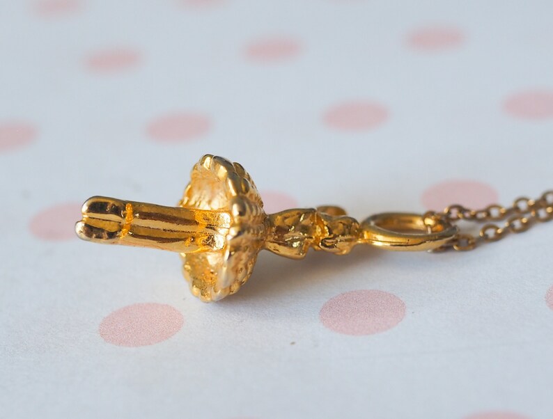 May include: A gold-colored pendant necklace featuring a detailed ballerina figure. The ballerina is in a classical pose, with arms extended. The necklace chain is delicate and complements the pendant's intricate design. The background is a soft, blurred pattern.
