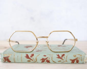 May include: Gold-framed eyeglasses with a geometric design rest on a vintage book. The book has a light green cover with floral and leaf patterns in red and gold. The eyeglasses have a classic, retro style.