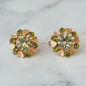 Peut inclure: Une paire de boucles d'oreilles en or avec un centre en strass vert entour&eacute; de petits strass verts et d'accents en feuilles d'or.