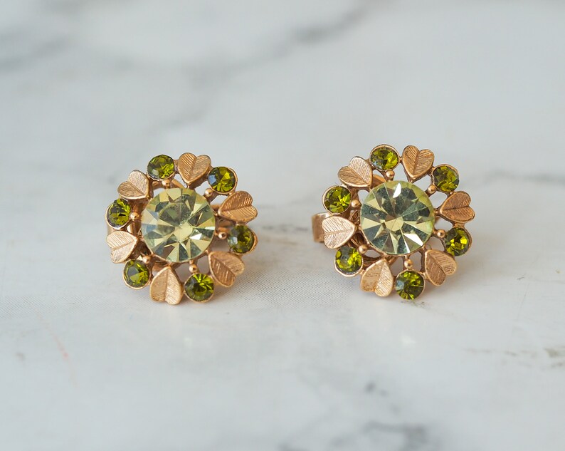 Peut inclure: Une paire de boucles d'oreilles en or avec un centre en strass vert entour&eacute; de petits strass verts et d'accents en feuilles d'or.