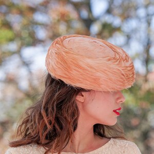 May include: A woman wearing a peach-colored feathered hat. The hat is large and covers most of her forehead. She is wearing a white lace dress.