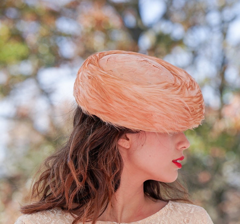 May include: A woman wearing a peach-colored feathered hat. The hat is large and covers most of her forehead. She is wearing a white lace dress.