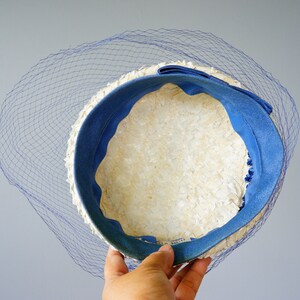May include: A vintage hat with a cream-coloured woven design, a vibrant blue interior, and a matching blue bow. A delicate blue netting veil drapes over the hat's brim, adding a touch of elegance. The hat is held by a hand.