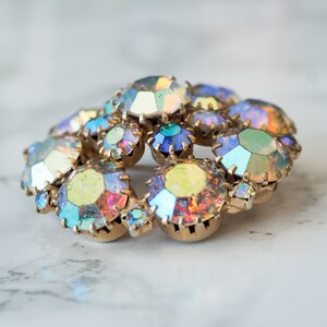 May include: A cluster of iridescent rhinestones in various shades of blue, green, and yellow, set in a gold tone metal base. The rhinestones are arranged in a circular pattern, creating a sparkling and eye-catching design.