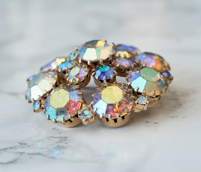 May include: A cluster of iridescent rhinestones in various shades of blue, green, and yellow, set in a gold tone metal base. The rhinestones are arranged in a circular pattern, creating a sparkling and eye-catching design.