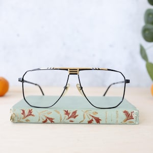Könnte beinhalten: Geometrische Brille mit schwarzem und goldenem Rahmen und goldenem Steg, die auf einem Vintage-Buch mit floralem Muster liegt. Zwei Orangen und eine Pflanze befinden sich im Hintergrund. Die Brille hat einen schwarzen Rahmen.