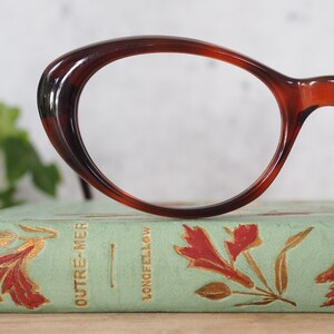 May include: A pair of tortoiseshell cat-eye glasses resting on a vintage book with gold lettering that reads "OUTRE-MER" and "LONGFELLOW".