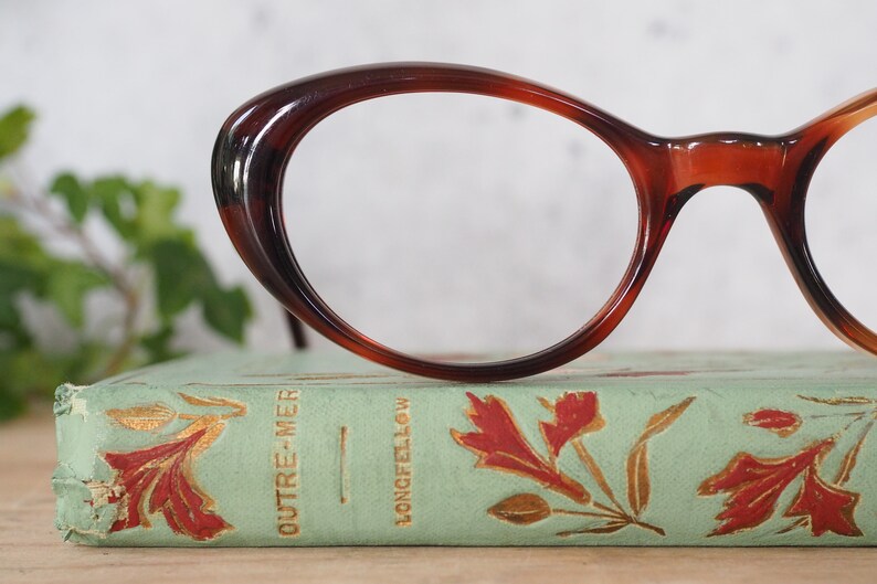 May include: A pair of tortoiseshell cat-eye glasses resting on a vintage book with gold lettering that reads "OUTRE-MER" and "LONGFELLOW".