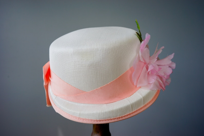 Op de afbeelding: Een witte hoed met een perzikkleurig lint en een roze bloem.