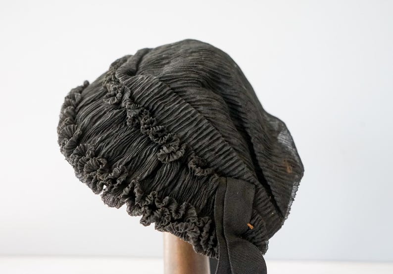 May include: A black, ruffled, fabric hat with a black ribbon. The hat is made of a soft, sheer material and has a delicate, feminine look.