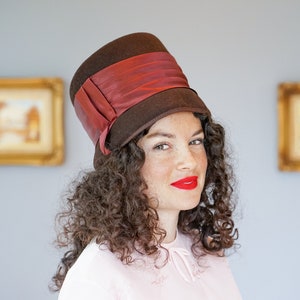 May include: A woman with curly brown hair is wearing a brown felt hat with a wide red ribbon. She is wearing a light pink blouse and has red lipstick on.