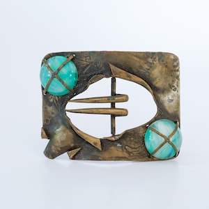 May include: A square, vintage belt buckle with a bronze finish. The buckle features a central oval cutout and two turquoise-colored, spherical stones. The design includes three bronze prongs and a decorative, textured surface.