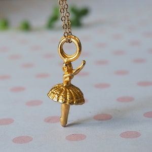 May include: A gold-colored pendant necklace featuring a ballerina charm. The charm depicts a ballerina in a tutu with one arm raised. The necklace chain and charm are a shiny gold color. The background is a soft, blurred pattern.