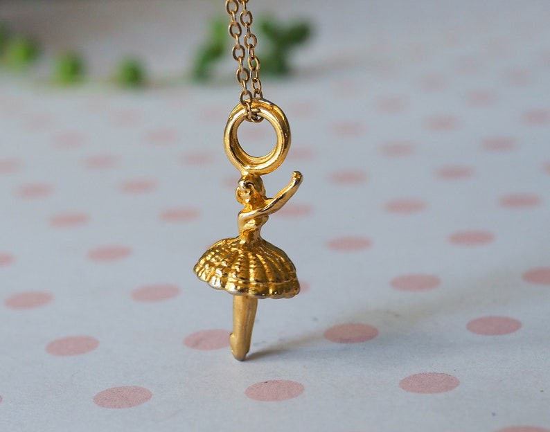 May include: A gold-colored pendant necklace featuring a ballerina charm. The charm depicts a ballerina in a tutu with one arm raised. The necklace chain and charm are a shiny gold color. The background is a soft, blurred pattern.