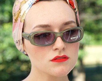 Könnte beinhalten: Olivgrüne rechteckige Sonnenbrille mit dunklen Gläsern. Die Sonnenbrille hat einen UV 400 Aufkleber. Die Person trägt ein gemustertes Kopftuch mit rosa Hintergrund und roten Lippenstift.