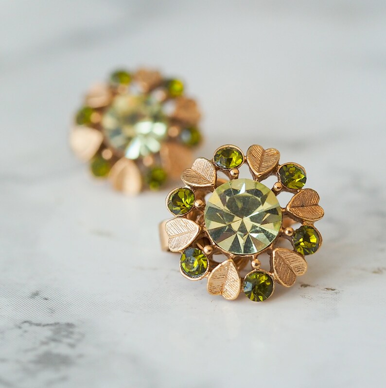 Peut inclure: Une paire de boucles d'oreilles en or avec un motif floral. Les boucles d'oreilles pr&eacute;sentent un grand strass vert au centre entour&eacute; de petits strass verts et de feuilles dor&eacute;es.