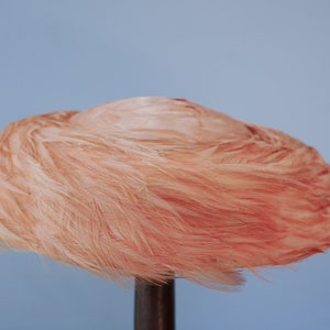 May include: A pink feather hat with a round shape. The hat is displayed on a wooden stand.