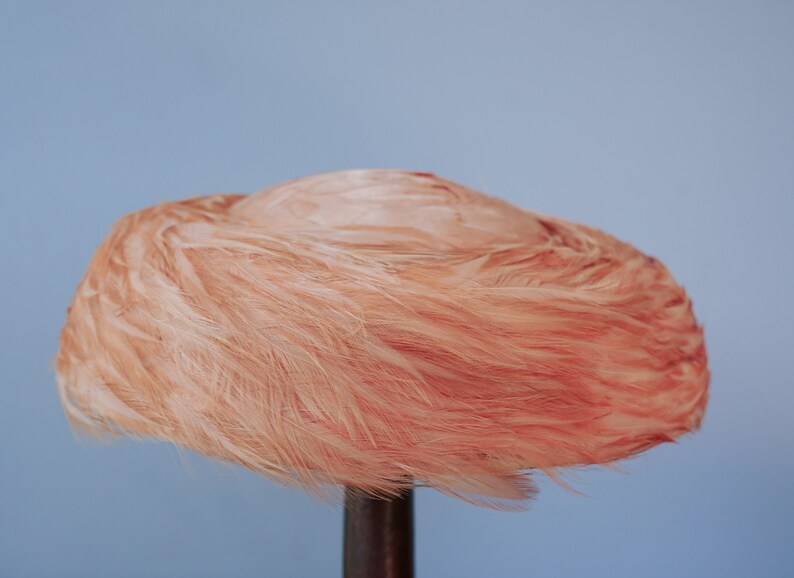 May include: A pink feather hat with a round shape. The hat is displayed on a wooden stand.
