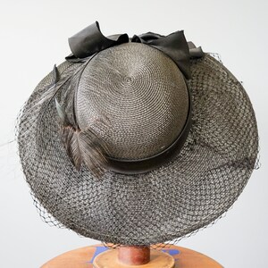 May include: A black straw hat with a black ribbon and a black feather. The hat has a wide brim and a net veil.