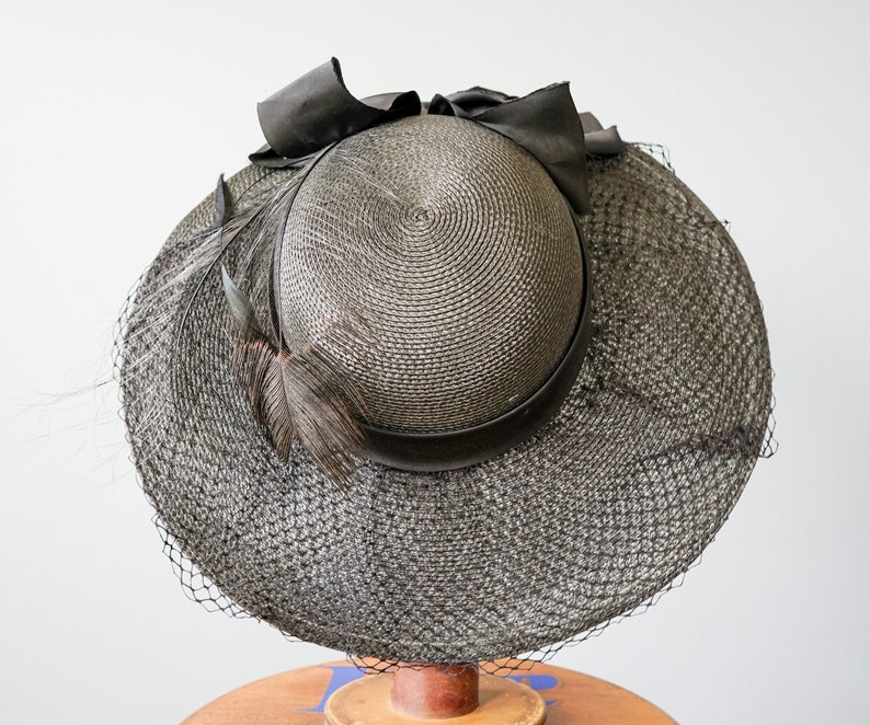 May include: A black straw hat with a black ribbon and a black feather. The hat has a wide brim and a net veil.