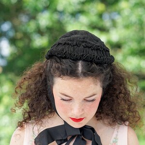 May include: A woman with curly brown hair wears a black lace bonnet with a ruffled edge. She is looking down with her eyes closed.
