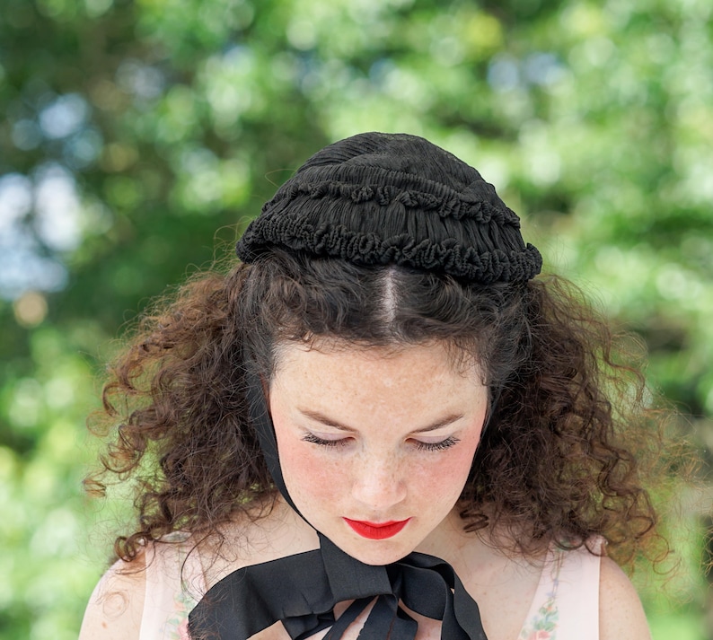 May include: A woman with curly brown hair wears a black lace bonnet with a ruffled edge. She is looking down with her eyes closed.